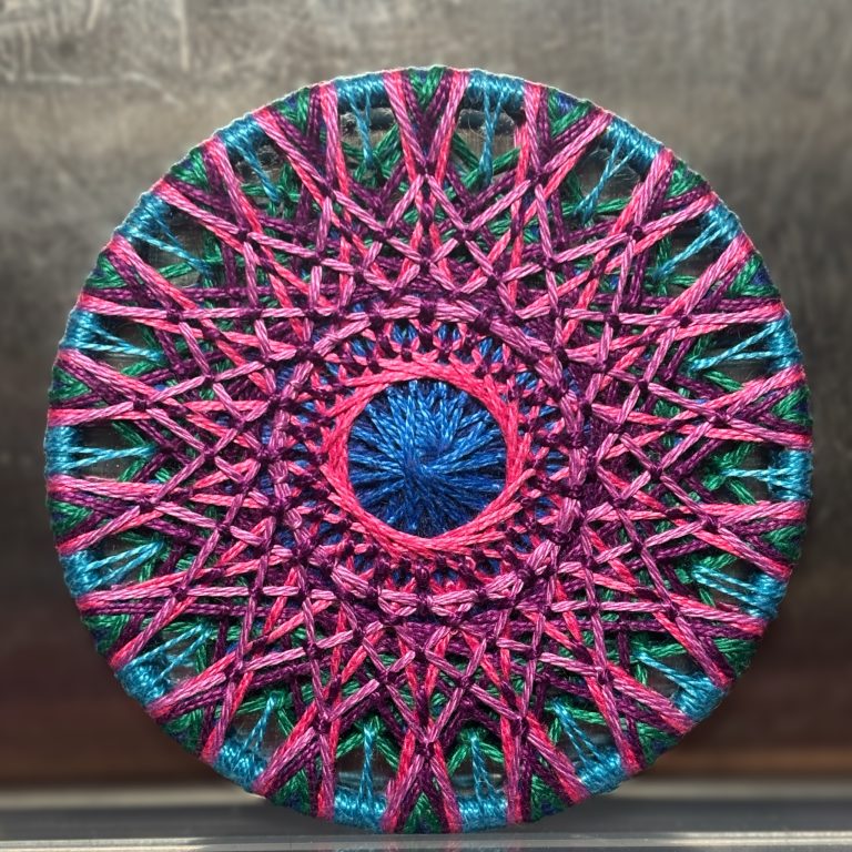 A 3" mandala made using zwirknopf button techniques. It uses navy, royal, and sky blues along with forest green, deep purple, hot pink, and mauve.