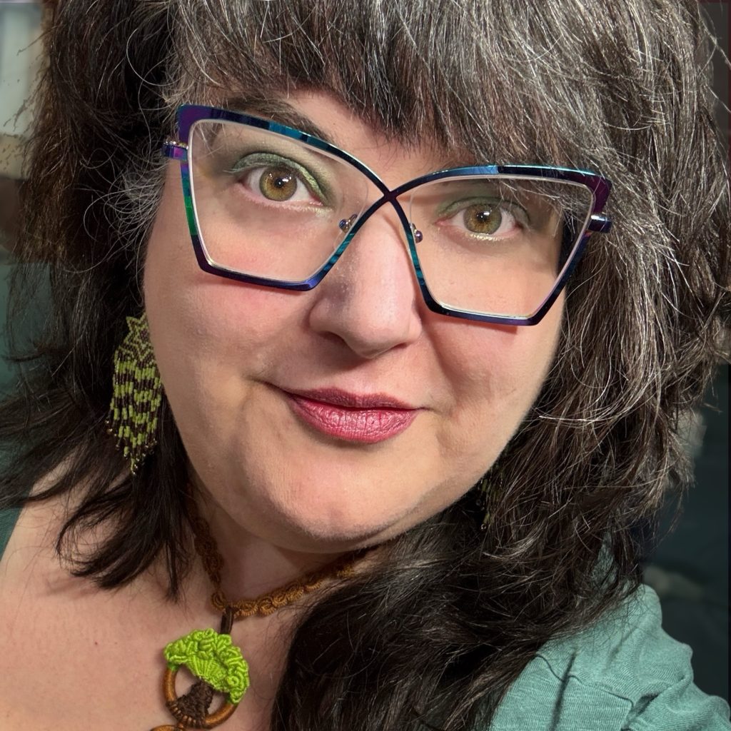 A woman with curly/wave brown hair with grey streaks is looking into the camera lens. She is wearing a green shirt, metallic rainbow glasses, handmade earrings with a Magen David on top and chevrons beneath, and a handmade Etz Chaim necklace.